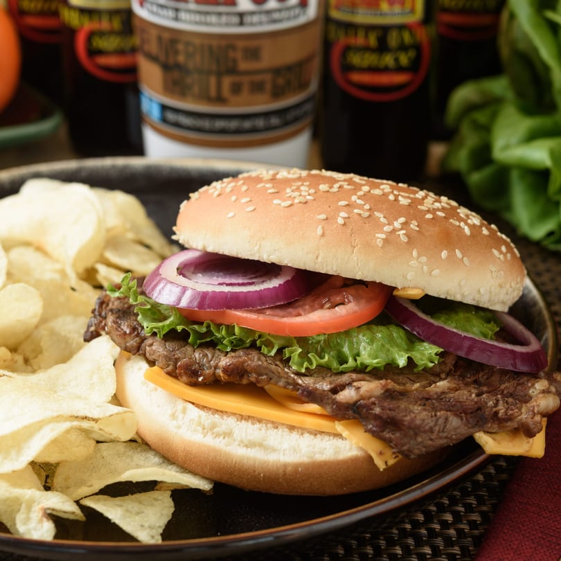 A steak sandwich with lettuce, tomato, red onion, and cheese, served with potato chips.